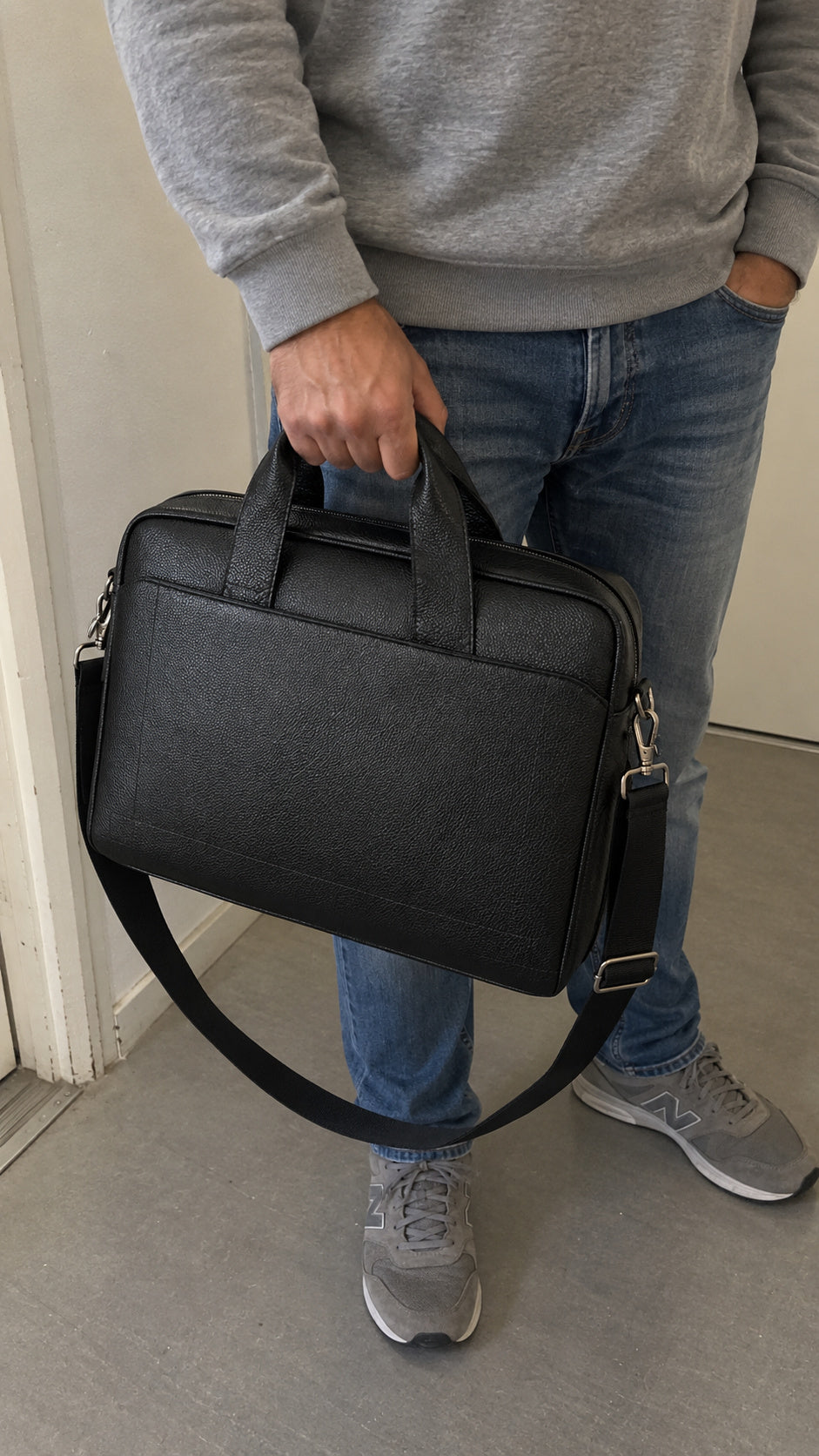 A person wearing a gray sweatshirt, blue jeans, and gray sneakers holds a black textured leather briefcase with a top handle and adjustable shoulder strap.