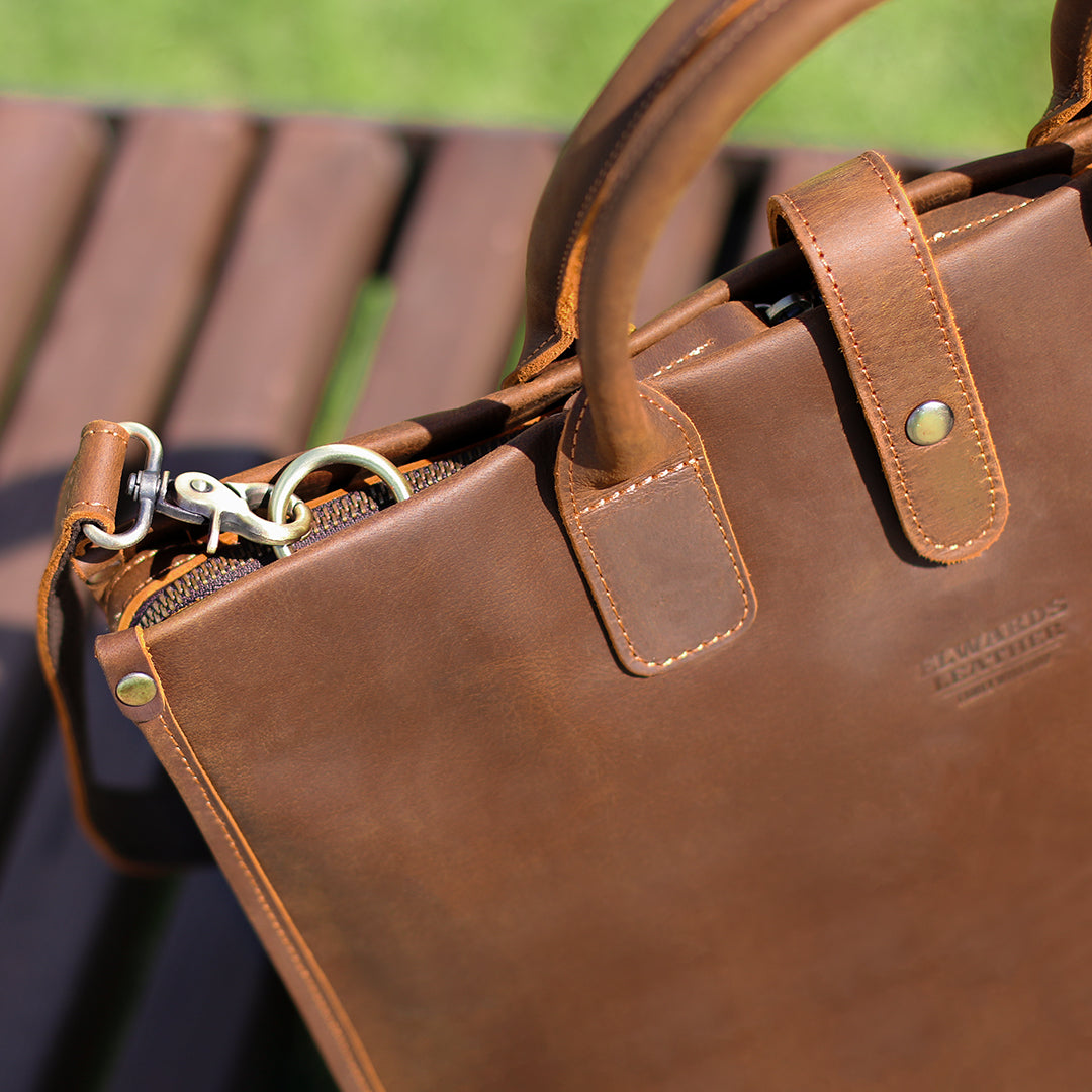 Close-up of brown men&#39;s Crazy Horse leather bag with reinforced handles and vintage metal details outdoors.