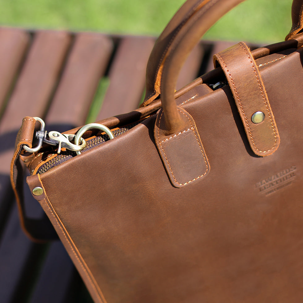 Close-up of brown men's Crazy Horse leather bag with reinforced handles and vintage metal details outdoors.