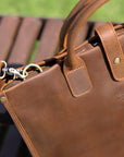 Close-up of brown men's Crazy Horse leather bag with reinforced handles and vintage metal details outdoors.