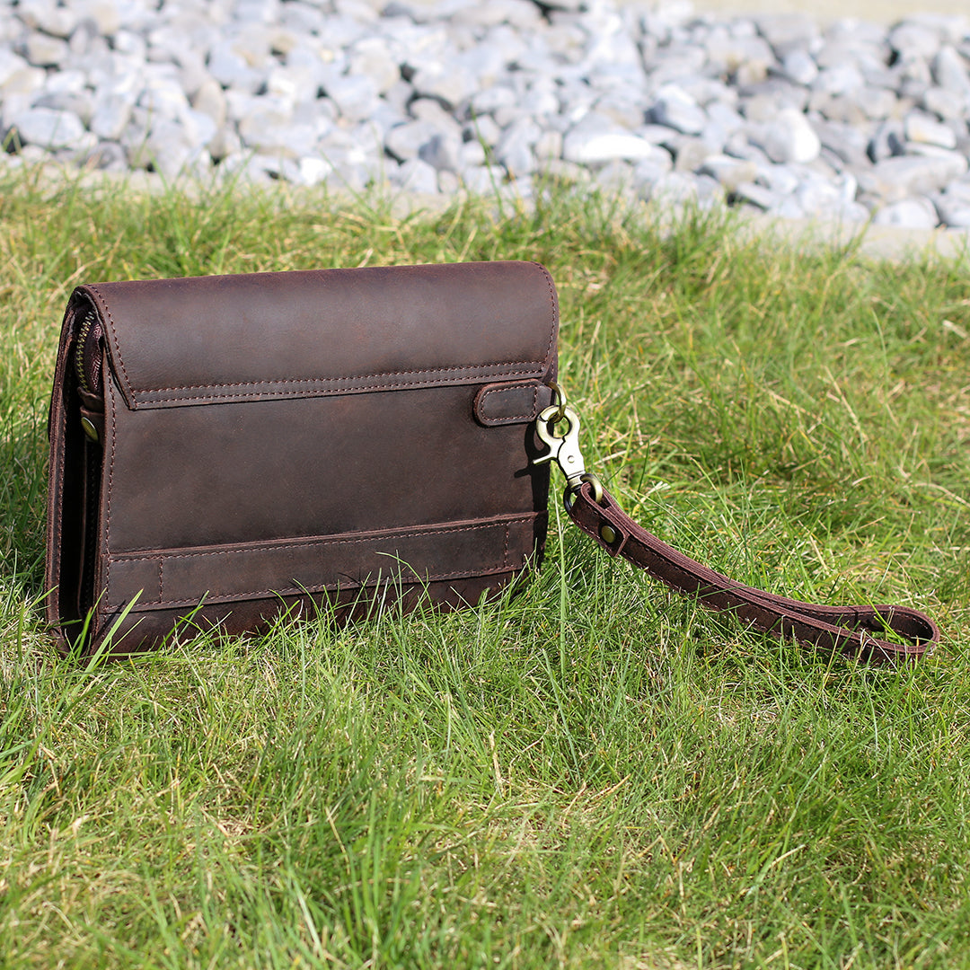 Back view of a dark brown leather clutch with a wrist strap on grass, highlighting its minimalist and practical style.