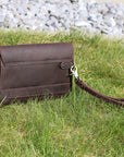 Back view of a dark brown leather clutch with a wrist strap on grass, highlighting its minimalist and practical style.