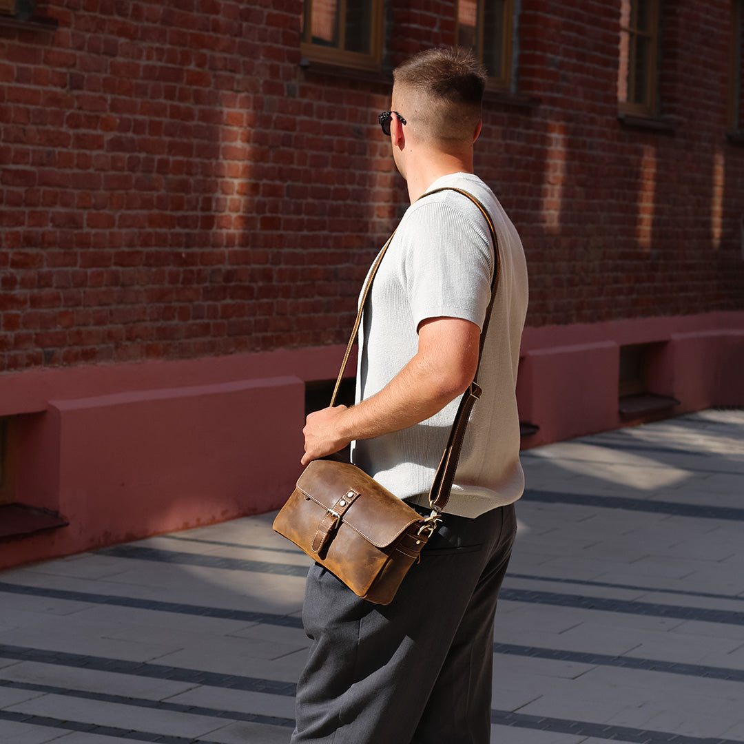 Man holding a brown leather bag against a brick wall.