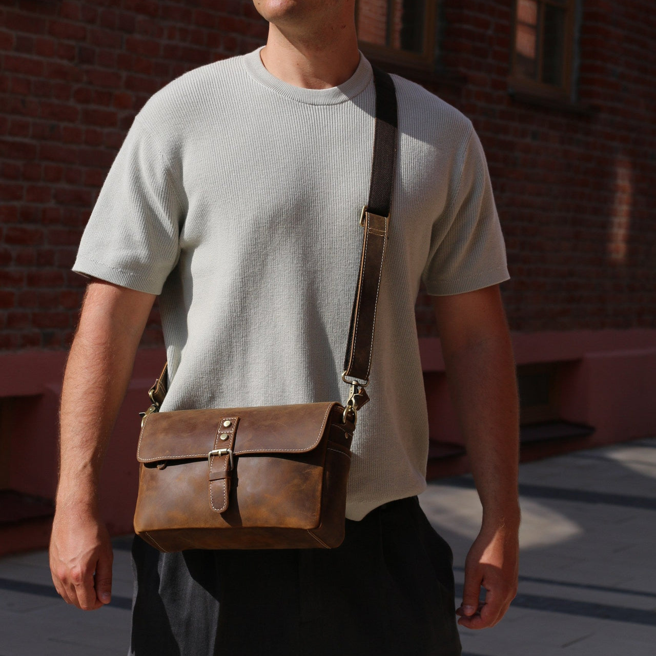 Man wearing sunglasses and a light gray t-shirt with a brown leather bag against a brick wall.