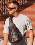 Man wearing a dark brown leather sling bag against a brick wall.