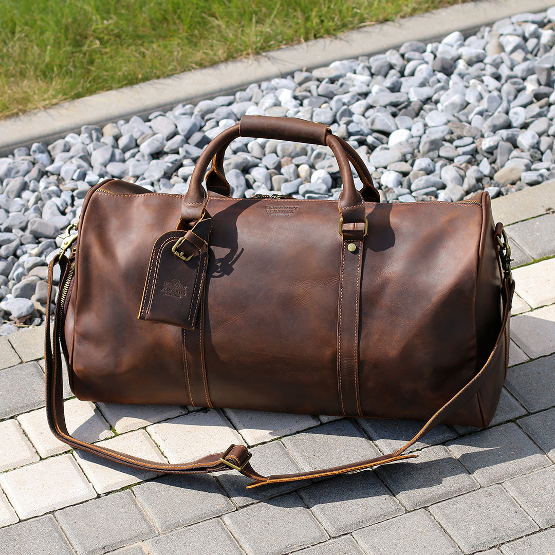 Full view of a handcrafted brown leather duffle bag with an adjustable strap, displayed outdoors.