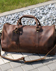 Full view of a handcrafted brown leather duffle bag with an adjustable strap, displayed outdoors.