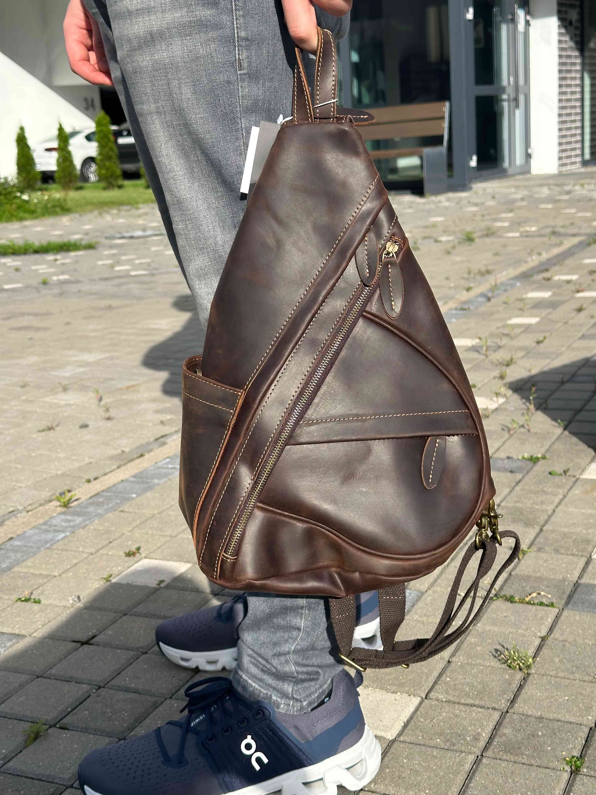 Brown leather sling bag worn by a person on a paved walkway.