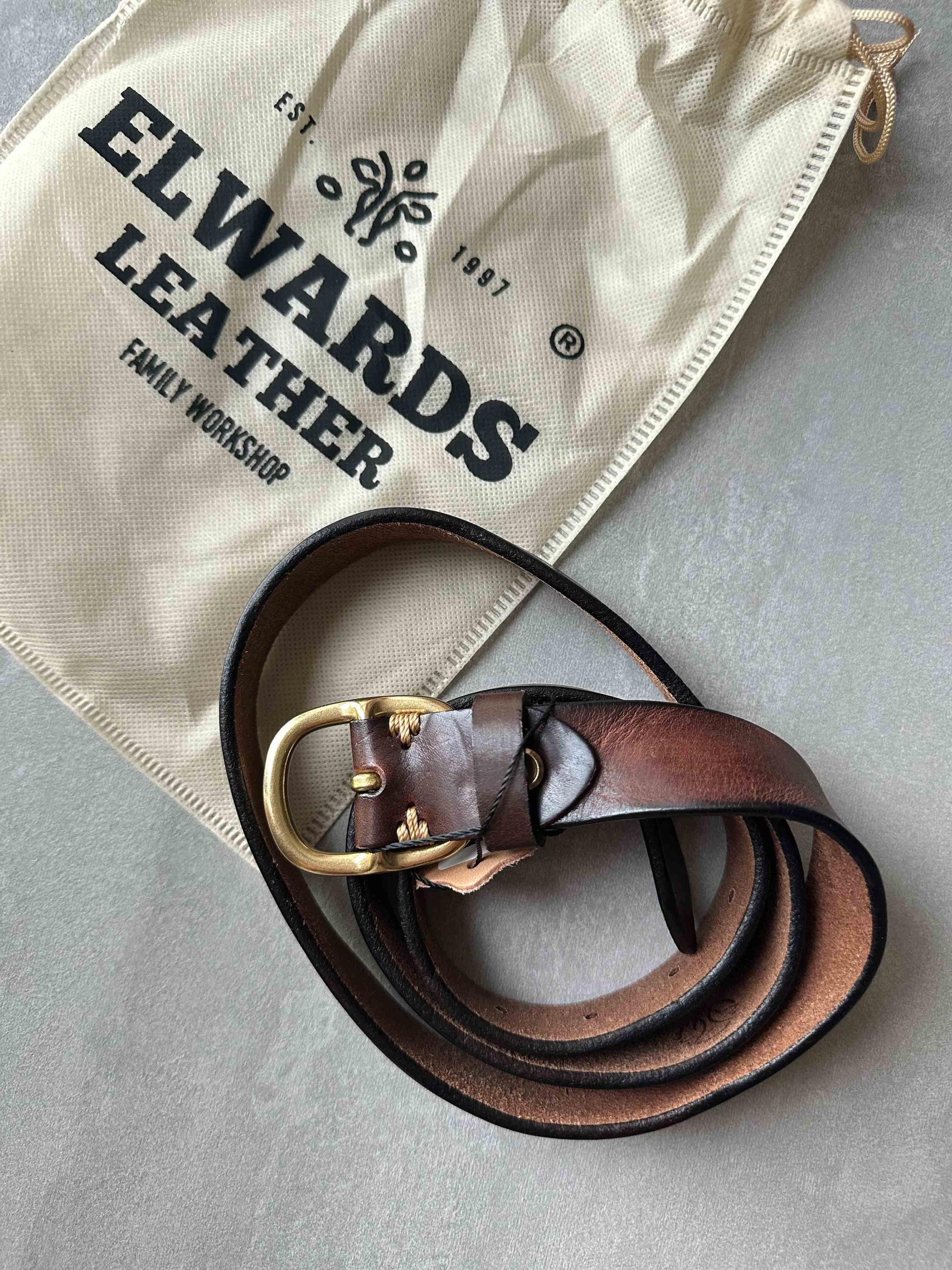Brown leather belt with gold buckle on a beige drawstring bag labeled 'Elwards Leather'.