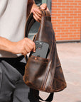 Person holding a brown leather sling bag with a phone case in an urban setting.