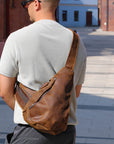 Man wearing a brown leather sling bag outdoors on a sunny day.
