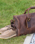 Handmade brown leather duffle bag with dedicated shoe compartment, shown outdoors with casual shoes partially stored.