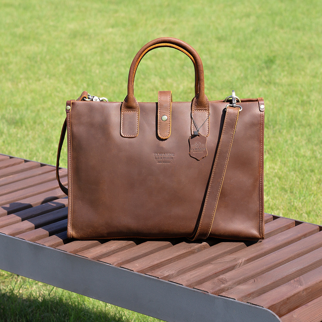 Handcrafted brown laptop leather briefcase resting on a bench in a sunny park.