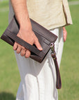 Side view of a men's dark brown clutch being held by its sturdy grip strap, illustrating its ergonomic and functional design.