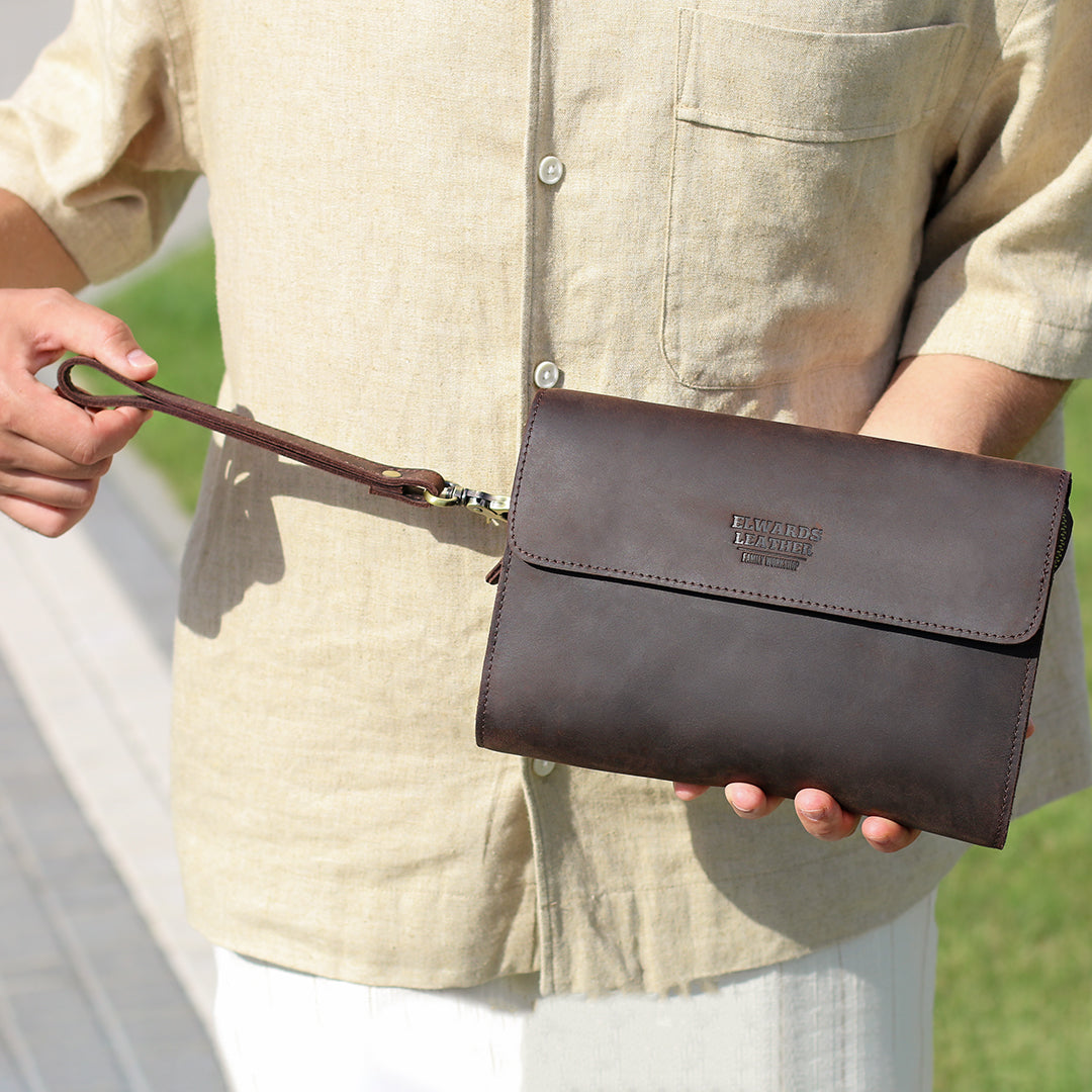 A man holding a dark brown clutch with a wrist strap outdoors, showcasing the versatility and premium look of the accessory.