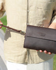 A man holding a dark brown clutch with a wrist strap outdoors, showcasing the versatility and premium look of the accessory.