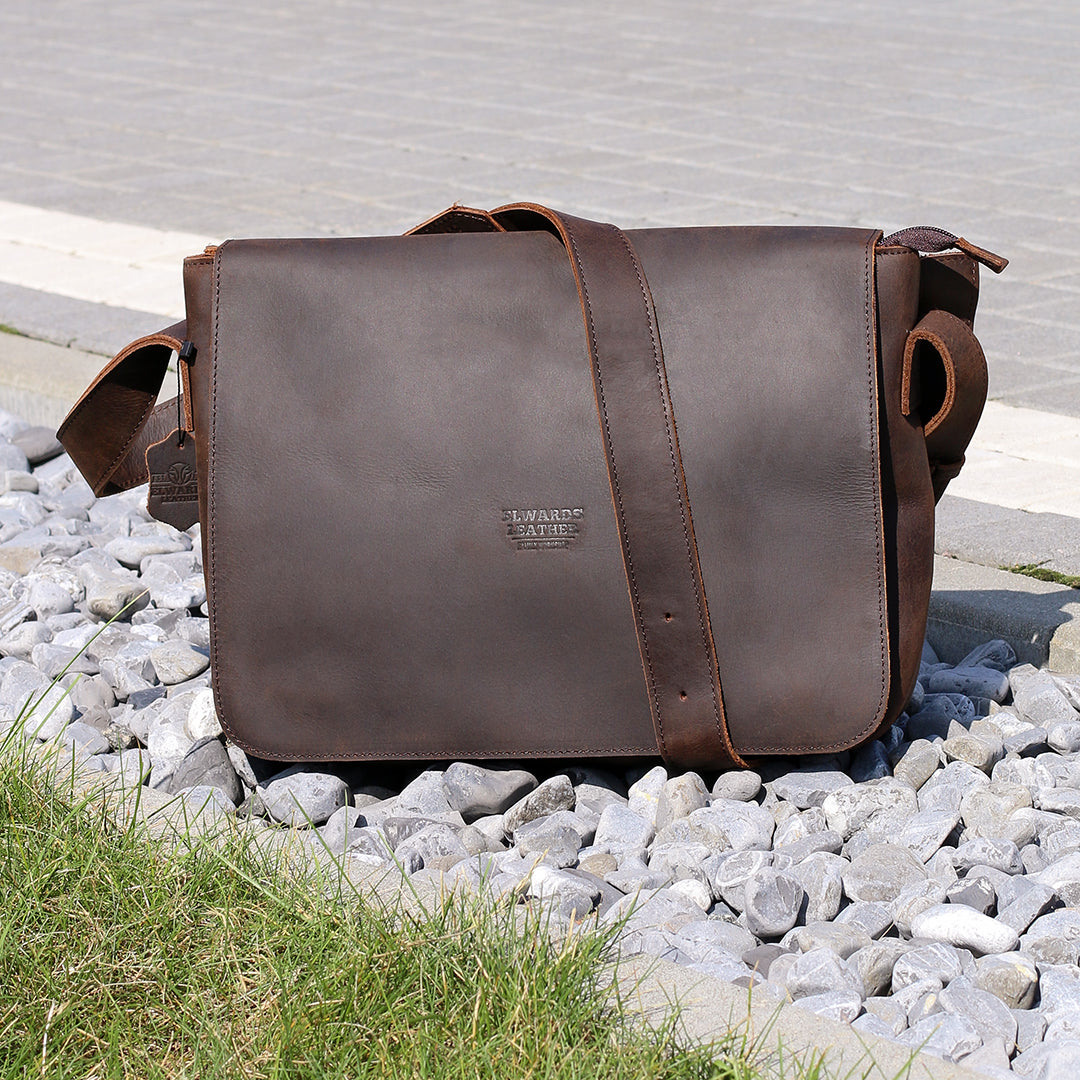 Dark brown leather messenger bag displayed on gravel, highlighting its sleek design and durability