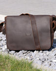 Dark brown leather messenger bag displayed on gravel, highlighting its sleek design and durability