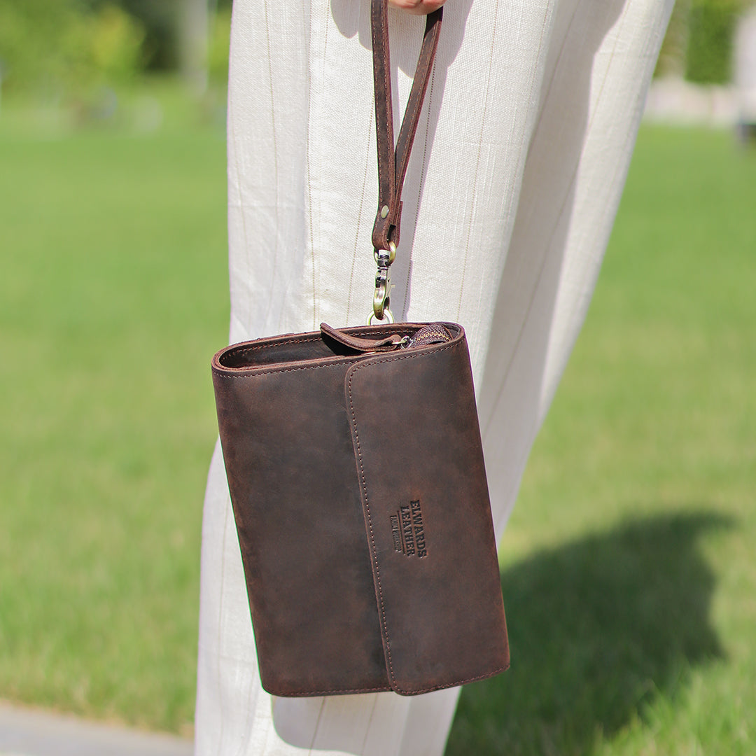 Stylish dark brown leather wrist bag being carried by hand, highlighting its compact design and versatility for outdoor use.