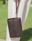 Stylish dark brown leather wrist bag being carried by hand, highlighting its compact design and versatility for outdoor use.