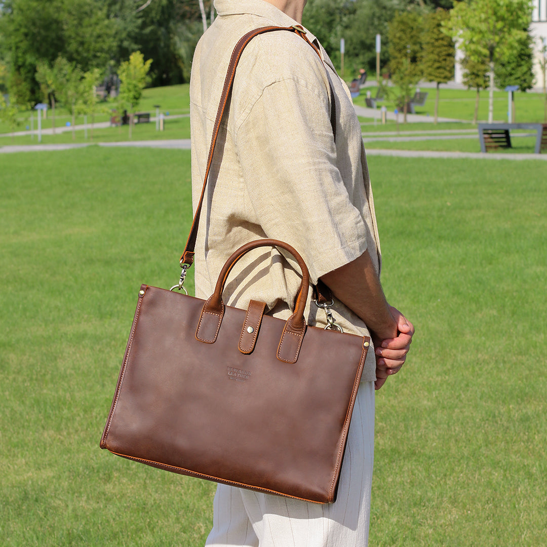 Man carrying a handmade brown leather laptop shoulder bag with an adjustable strap in a park.