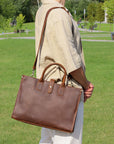 Man carrying a handmade brown leather laptop shoulder bag with an adjustable strap in a park.