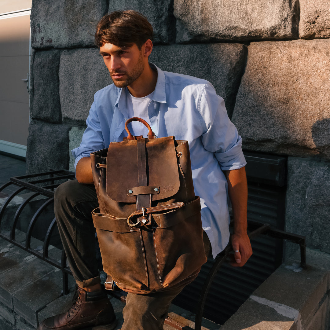 Man sitting outdoors with a vintage brown Crazy Horse leather backpack, highlighting its durability and compartments.