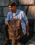 Man sitting outdoors with a vintage brown Crazy Horse leather backpack, highlighting its durability and compartments.