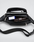 Black leather crossbody bag shown open from above, with multiple zip compartments and a wide adjustable strap. A white smartphone, brown wallet, and small blue item are visible inside on a light gray studio background.