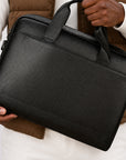 A black pebbled leather laptop briefcase with top handles, held by a person wearing a brown puffer vest and white long-sleeve shirt.