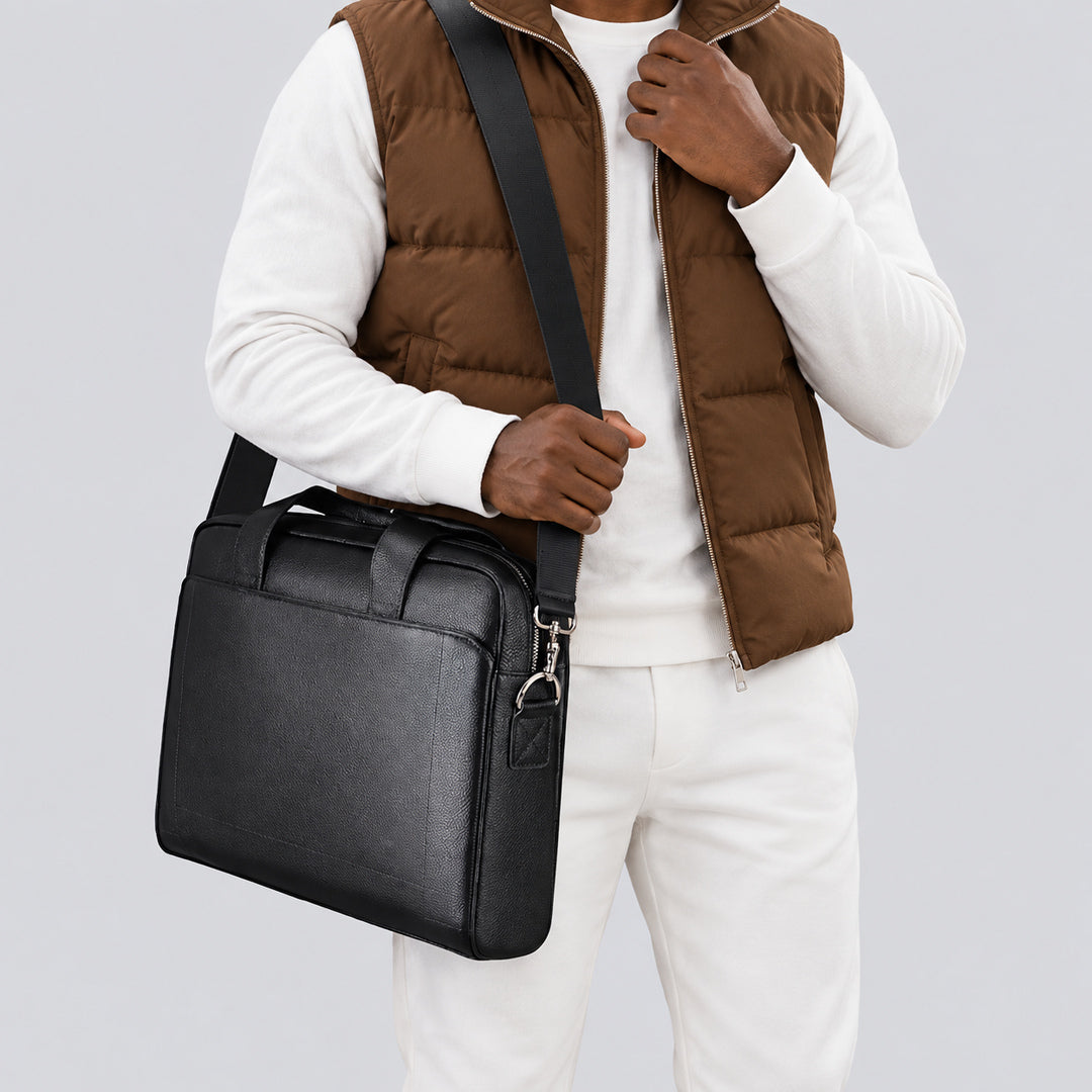 A man wearing a brown puffer vest, white long-sleeve shirt, and white pants carries a black leather messenger bag with a wide adjustable shoulder strap against a light gray studio background.