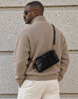 Back view of a man wearing a black leather crossbody bag with gold-tone hardware