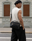 Man wearing a black leather crossbody bag at the hip, featuring a top handle, front zip pocket and adjustable strap.