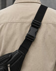 A close-up of a person wearing a beige jacket, showing a black leather crossbody bag with a wide black webbing strap and black plastic buckle.