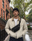 A man wearing a black beanie, cream jacket, and dark jeans stands on a wet city sidewalk wearing a black crossbody leather fanny pack. Brick townhouses and trees line the street in the background.