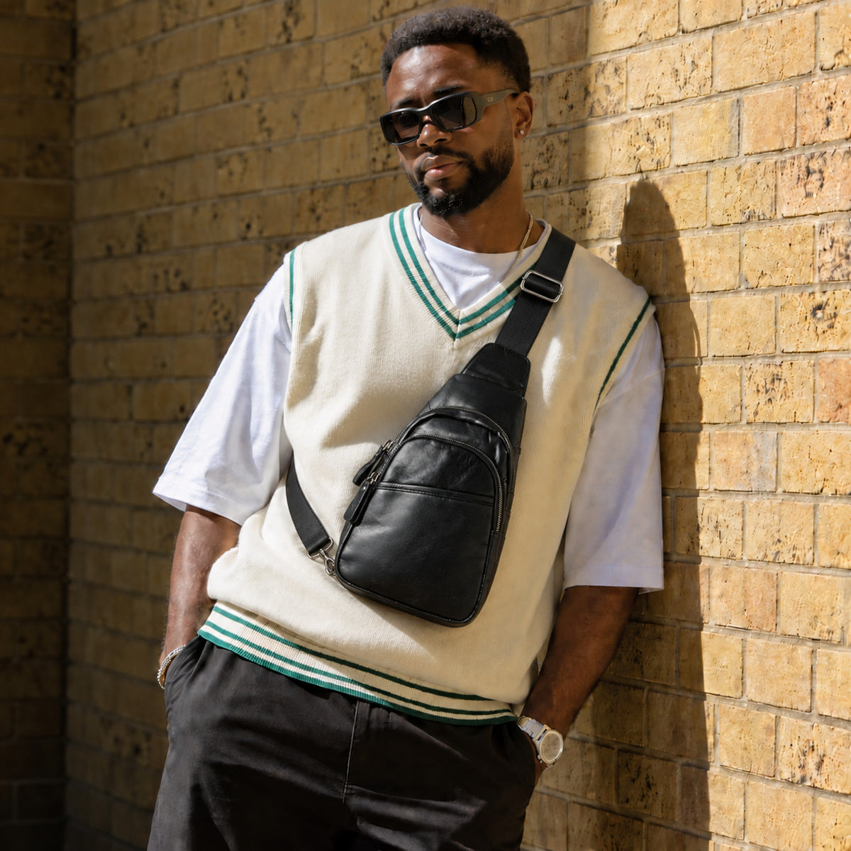 Man wearing a black leather crossbody sling bag with a cream sweater vest and sunglasses