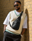 Man wearing a black leather crossbody sling bag with a cream sweater vest and sunglasses