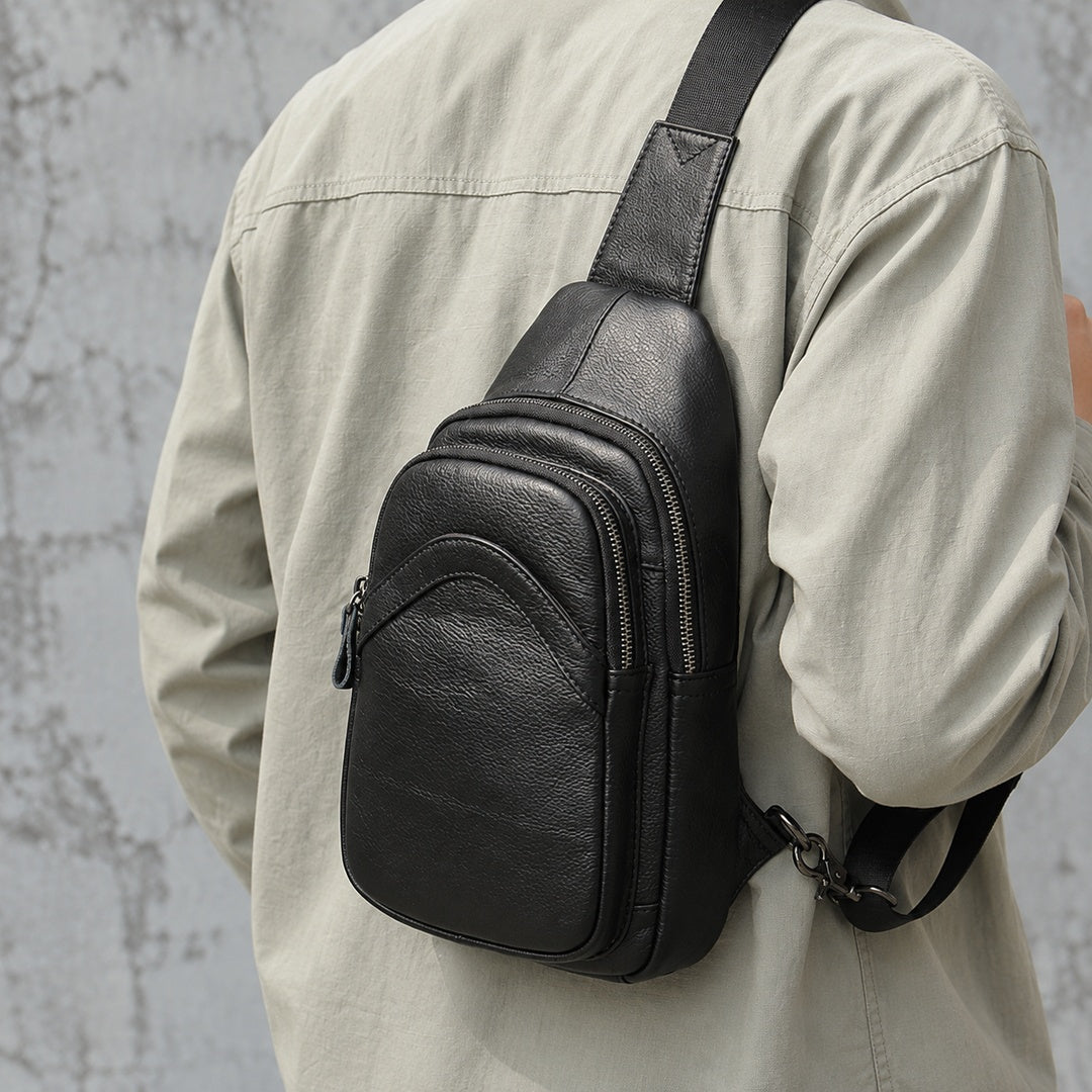 A black leather sling bag worn crossbody on a person's shoulder, shown from the back. The person is wearing a light beige button-up shirt against a textured gray concrete wall.