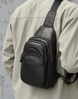 A black leather sling bag worn crossbody on a person's shoulder, shown from the back. The person is wearing a light beige button-up shirt against a textured gray concrete wall.