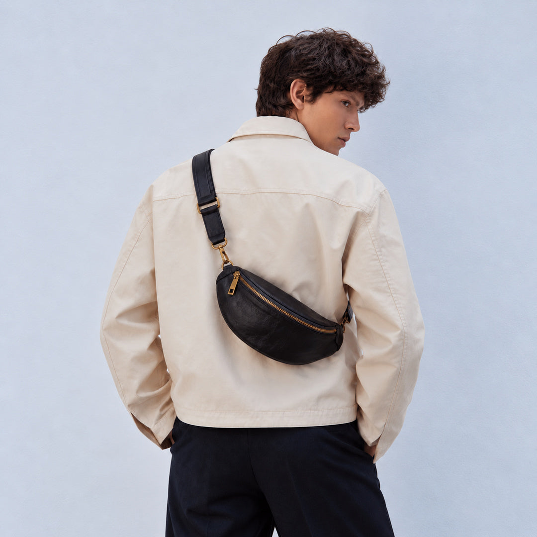 Back view of a man wearing a black leather sling bag crossbody with a wide adjustable strap and gold-tone hardware.