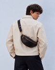 Back view of a man wearing a black leather sling bag crossbody with a wide adjustable strap and gold-tone hardware.