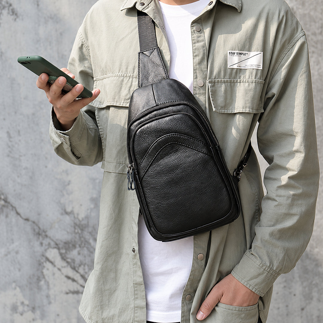 A man wearing a black leather crossbody phone bag across his chest, styled with an olive green shirt over a white t-shirt, holding a green smartphone against a gray concrete wall.