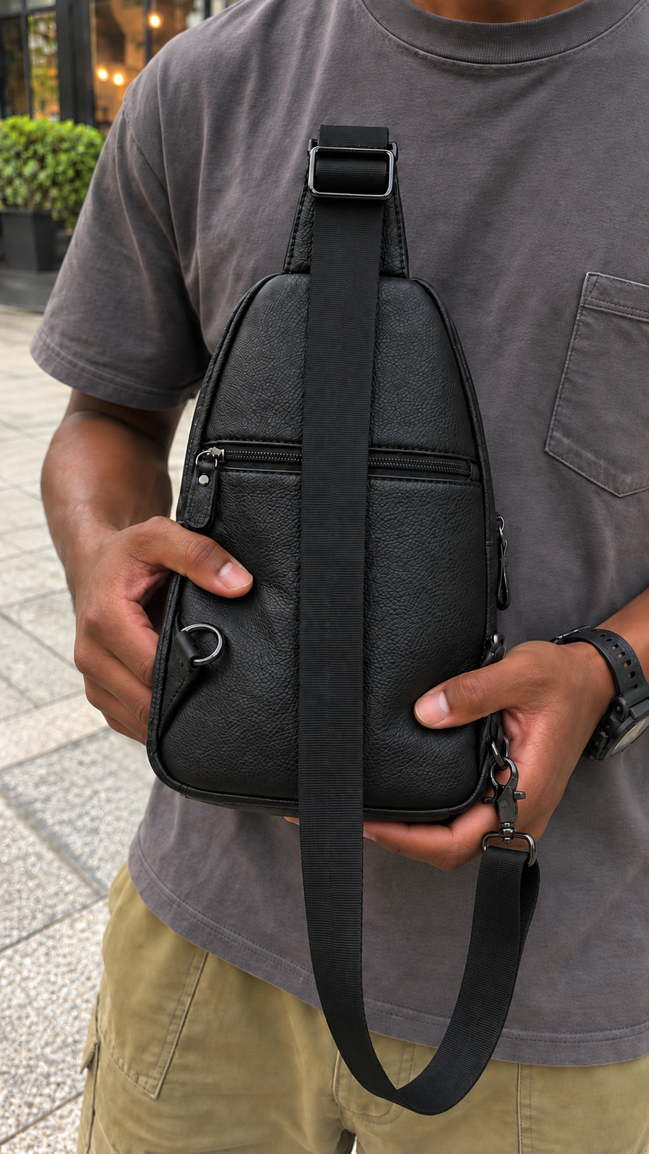 A person wearing a gray t-shirt and khaki pants holds a black textured leather crossbody bag with a wide adjustable strap. The bag features a front zippered pocket and a side ring detail, shown in an outdoor urban setting.