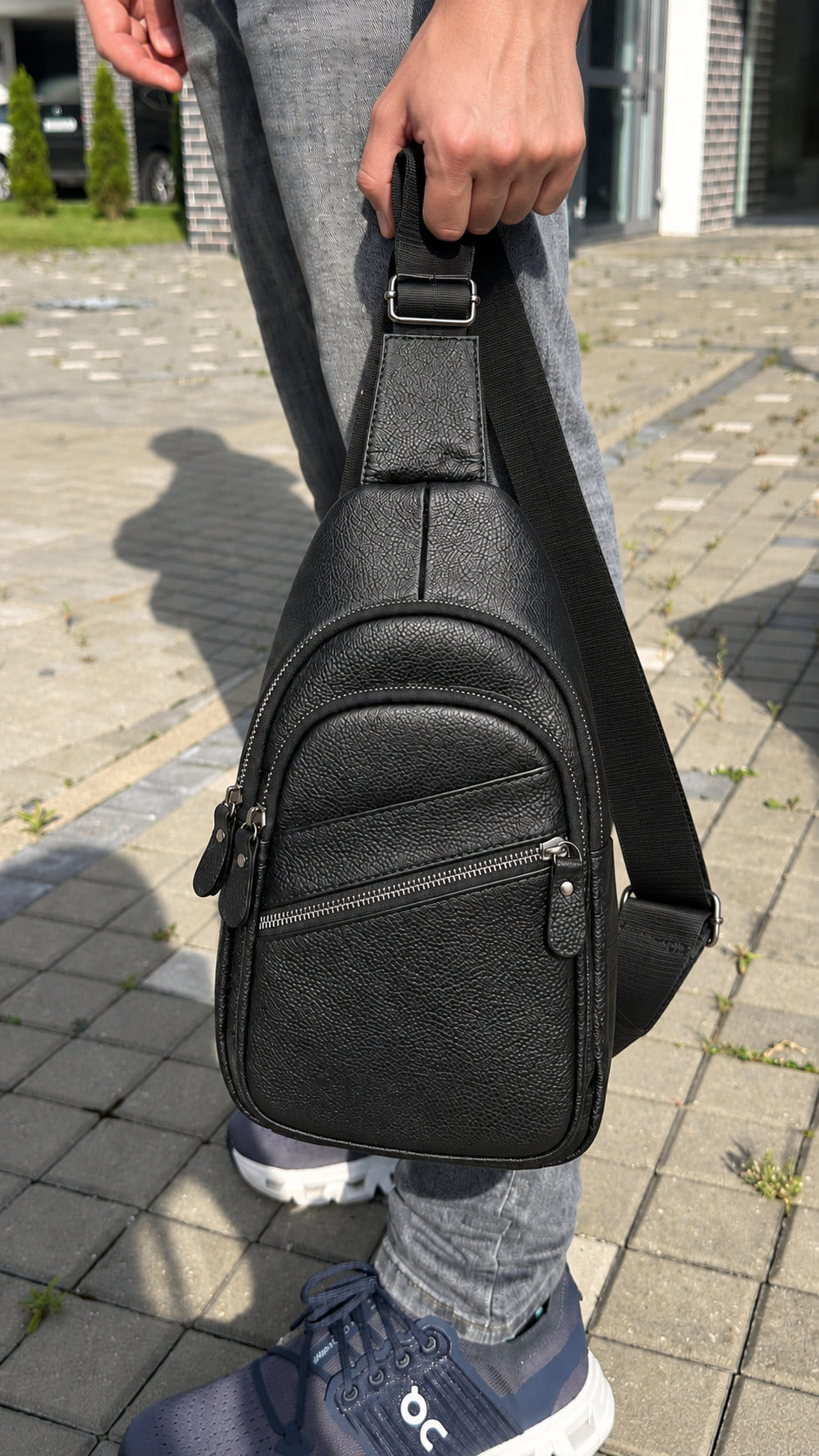 A black pebbled leather sling bag with an adjustable shoulder strap, held by a person wearing gray jeans and blue sneakers on a paved outdoor walkway.