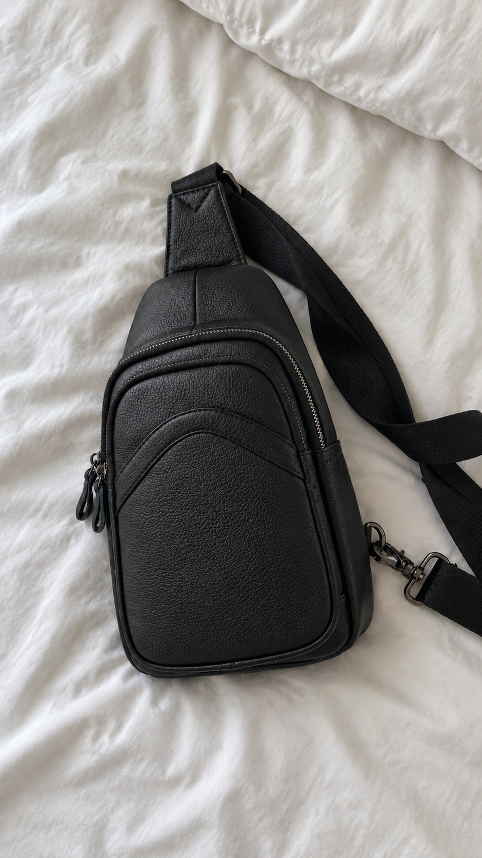 A black pebbled leather sling bag with a wide adjustable shoulder strap, laid flat on white textured bedding.