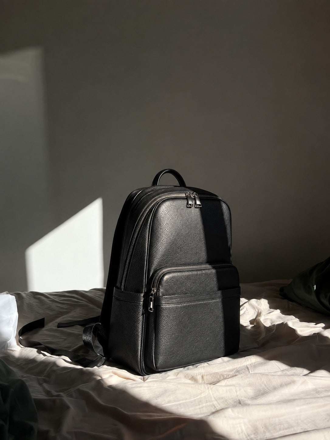 A black textured leather backpack sits on a bed with sunlight casting dramatic shadows on the wall and bedding.
