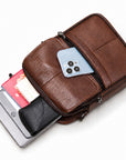 A brown textured leather crossbody bag shown open on a white background, with a light blue smartphone, a gray smartphone, cards, and a black cardholder partially visible inside.