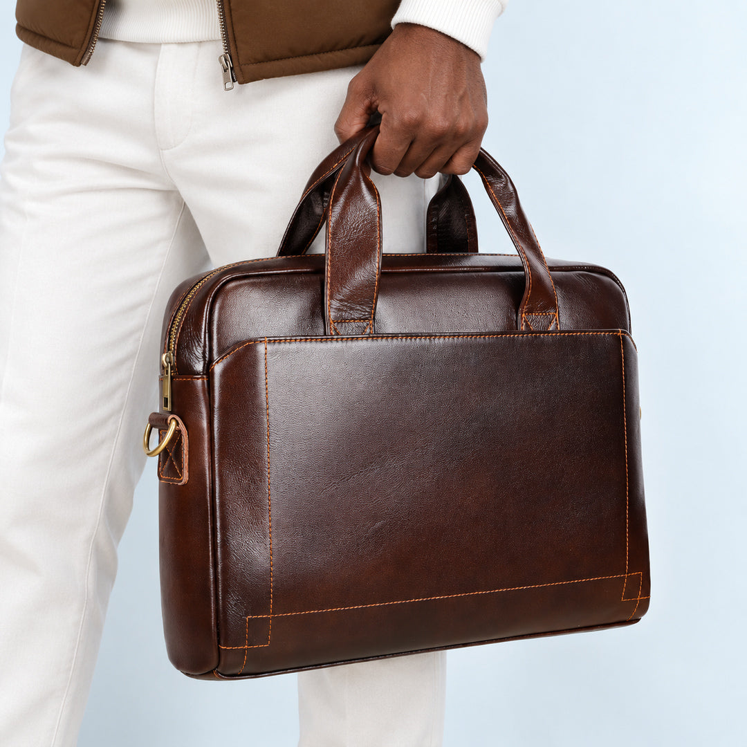 A man wearing white pants and a brown jacket holds a dark brown leather briefcase bag against a light blue background.