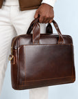 A man wearing white pants and a brown jacket holds a dark brown leather briefcase bag against a light blue background.