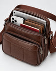 A brown textured leather crossbody bag with multiple compartments, shown open with a smartphone, wallet, and passport inside, on a light gray background.